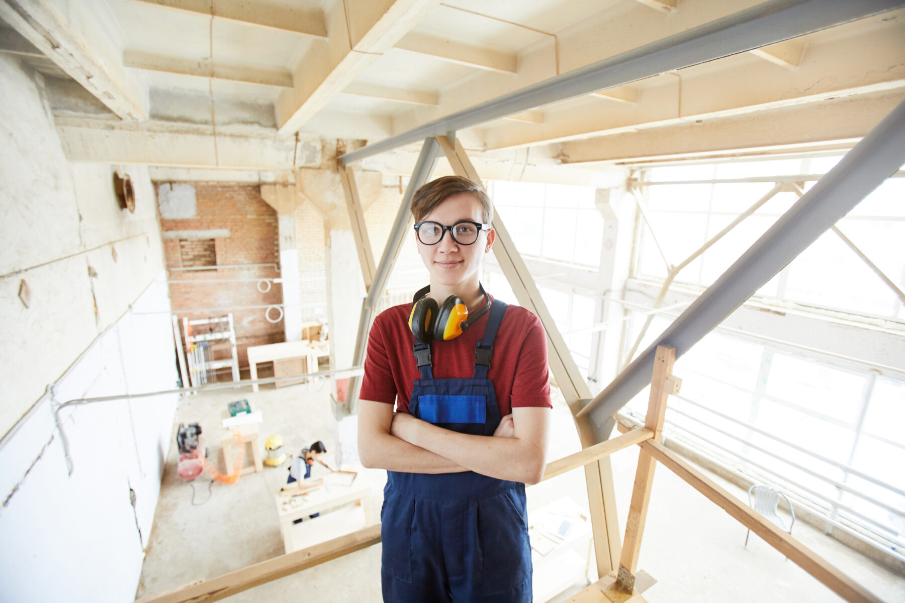 Content confident young carpenter in glasses wearing blue overall and yellow ear protectors on neck crossing arms on chest and looking at camera in workshop